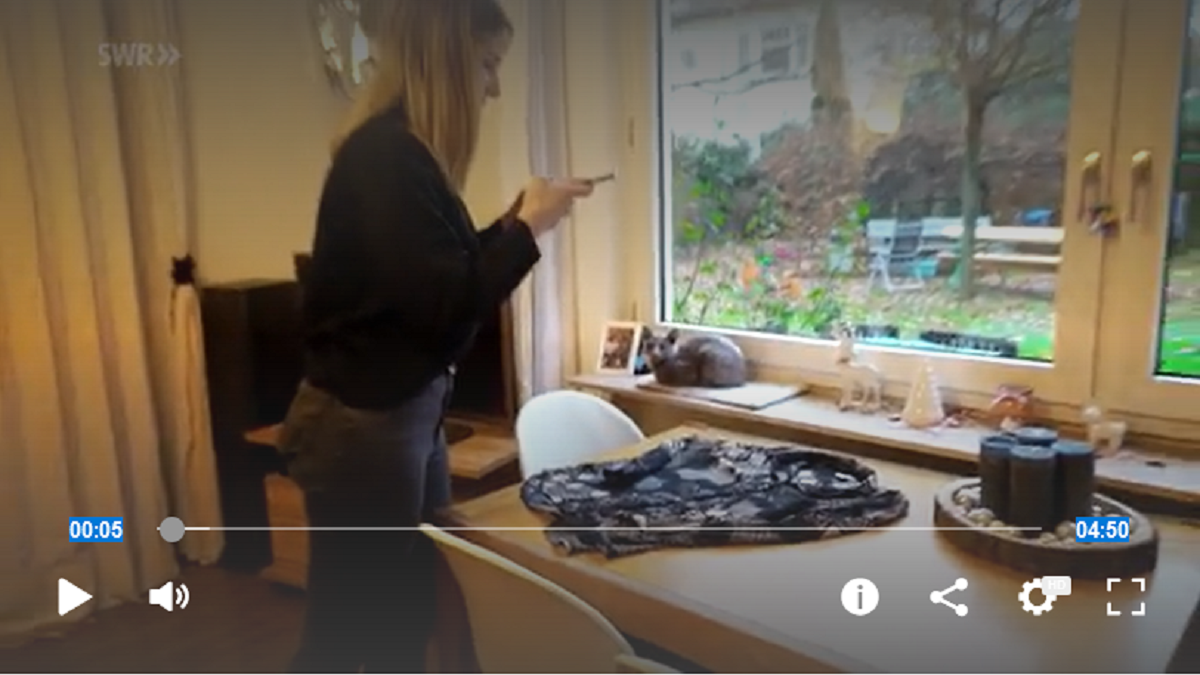 Ausschnitt eines Videos. Frau steht mit Smartphone am Fenster. Vor ihr steht ein Tisch.