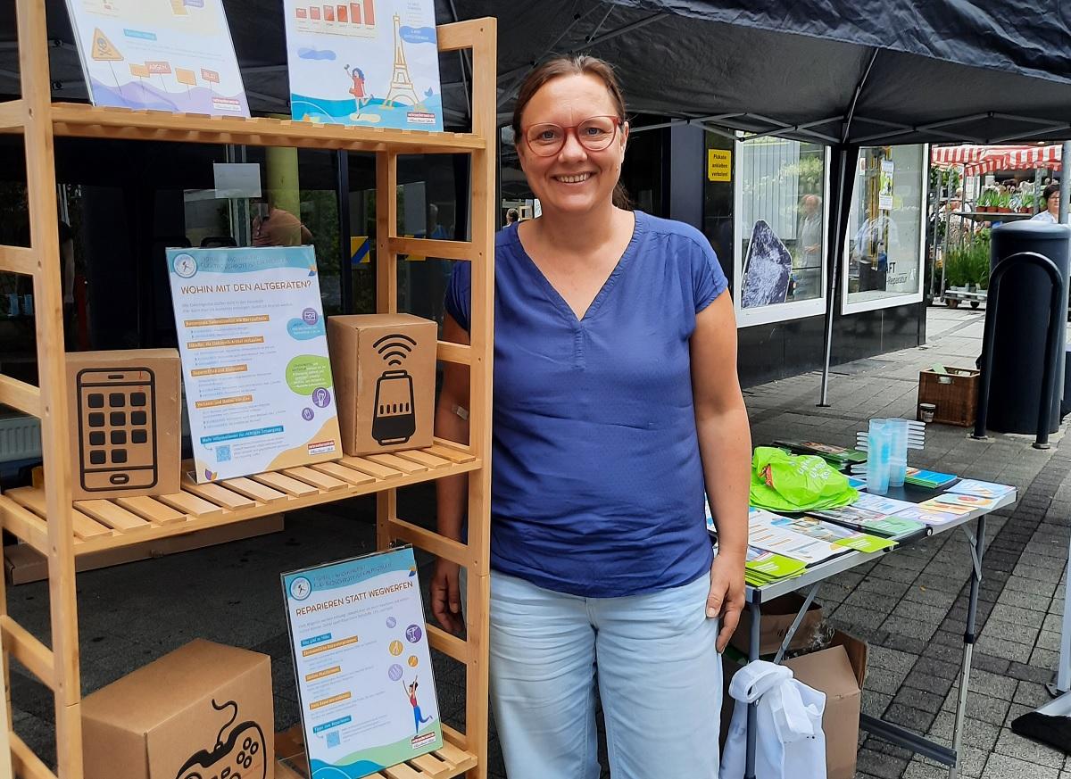 Eine Frau steht vor einem Regal mit Informationsmaterial und lächelt in die Kamera.