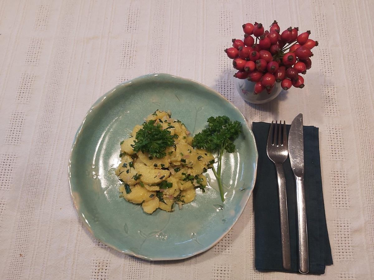 Eine Schale mit Kartoffelsalat mit Besteck. Daneben sthet noch eine Vase mit Hagebuttenzweigen.