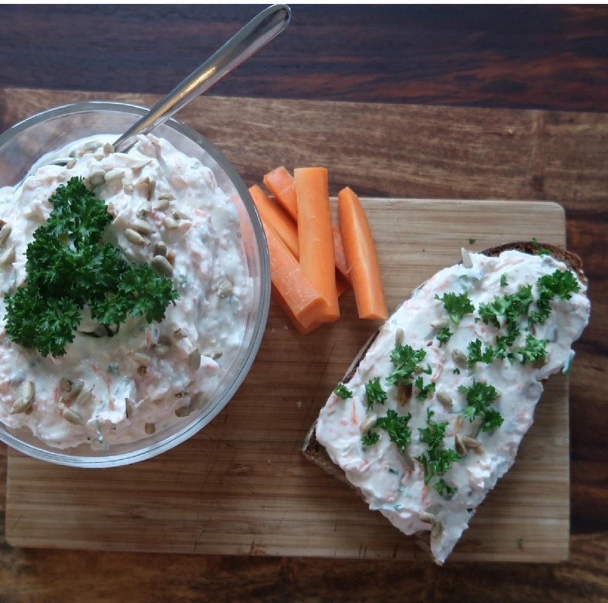 Eine Brotscheibe mit Quark bestrichen. Daneben steht noch eine Schüssel mit Quark und ein paar geschnittene Karotten liegen daneben.