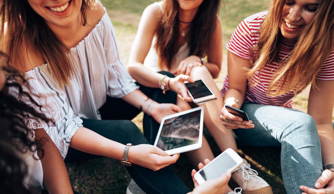 Eine Gruppe junger Frauen. Sie sitzen in einem Park und schauen auf ihre Handys und Tablets.