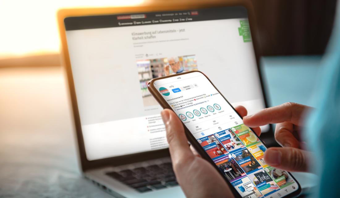 Ein Laptop auf einem Tisch. Davor eine Hand mit einem Smartphone in der Hand.