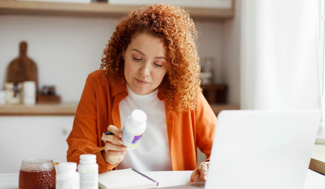 Eine rothaarige, lockige Frau sitzt am Küchentisch und schaut sich eine Packung Nahrungsergänzungsmittel an. Vor ihr steht ein Laptop.