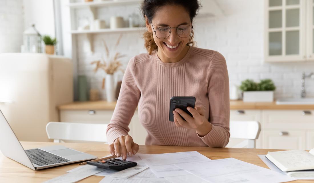 Frau sitzt am Schreibtisch mit vielen Rechnungen, sie schaut auf ihr Handy und tippt in den Taschenrechner
