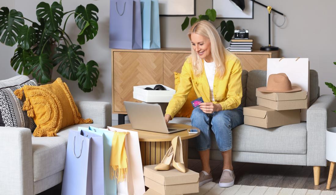 Eine Frau sitzt auf dem Sofa vor ihrem Laptop, in der Hand hält sie eine Kreditkarte. Sie lächelt, um sie herum sind viele Einkaufstüten verteilt, sowie ein Paar Highheels und ein Hut.