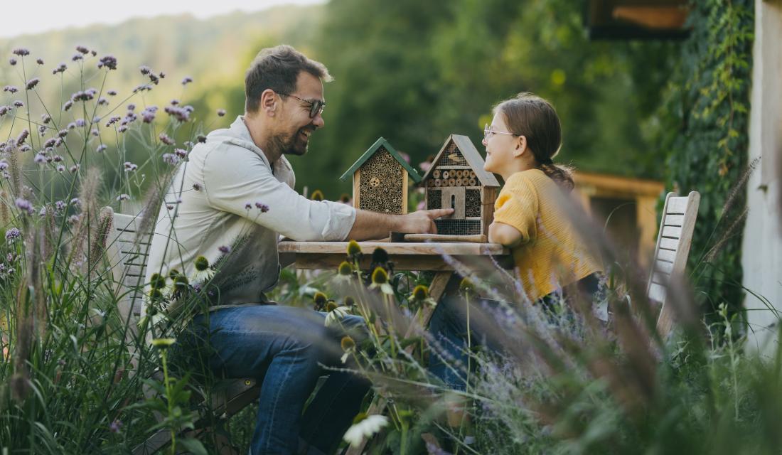 Vater und Tochter sitzen gemeinsam an einem Tisch im Garten umgeben von hohem Gras und Wildblumen und lachen sich an. Auf dem Tisch stehen zwei Insektenhäuser.