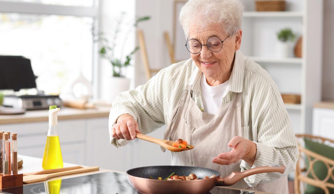 Alte Frau ist in der Küche und bereitet Gemüse in der Pfanne zu, sie lächelt dabei 