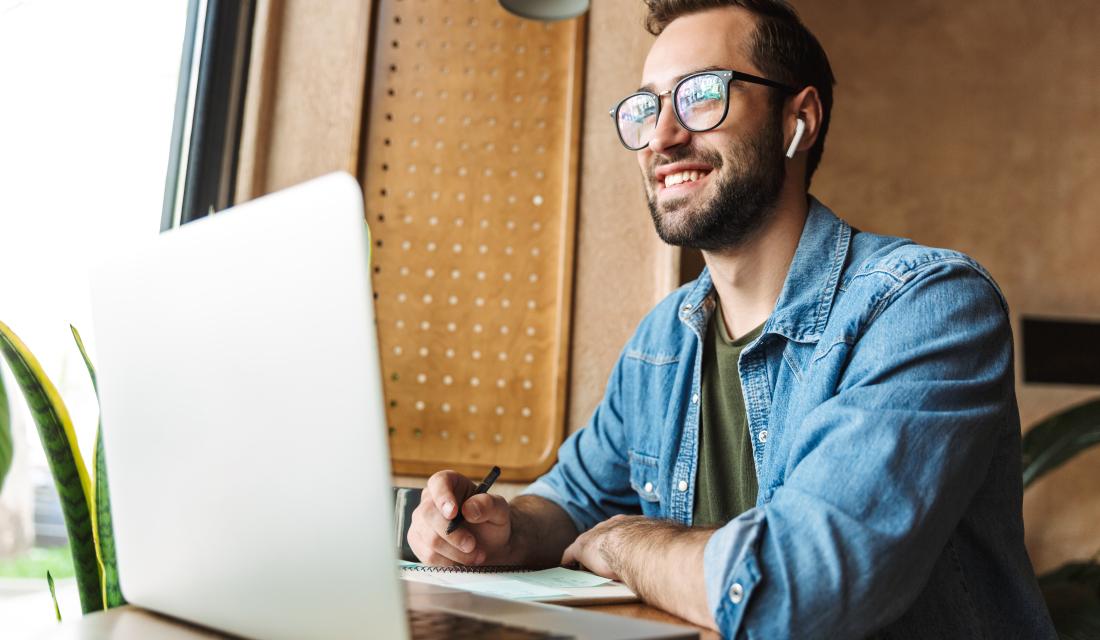 Junger Mann mit Brille sitzt in einem Café vor dem Laptop, er hält einen Stift in der rechten Hand und schreibt in einen Notizblock. Er blickt aus dem Fenster und lacht.