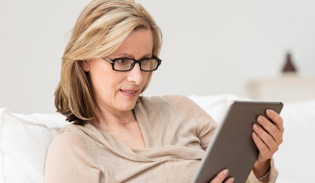 Frau mittleren Alters mit Brille sitzt auf einem Sofa. In ihren Händen hält sie ein silbernes Tablet, auf das sie schaut.
