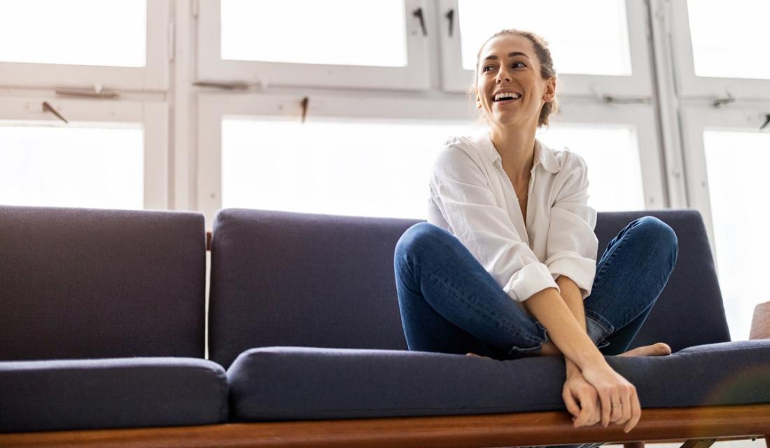 Eine Frau sitzt im Schneidersitz auf dem Sofa vor einer großen Fensterfront. Sie schaut zur Seite und lacht.