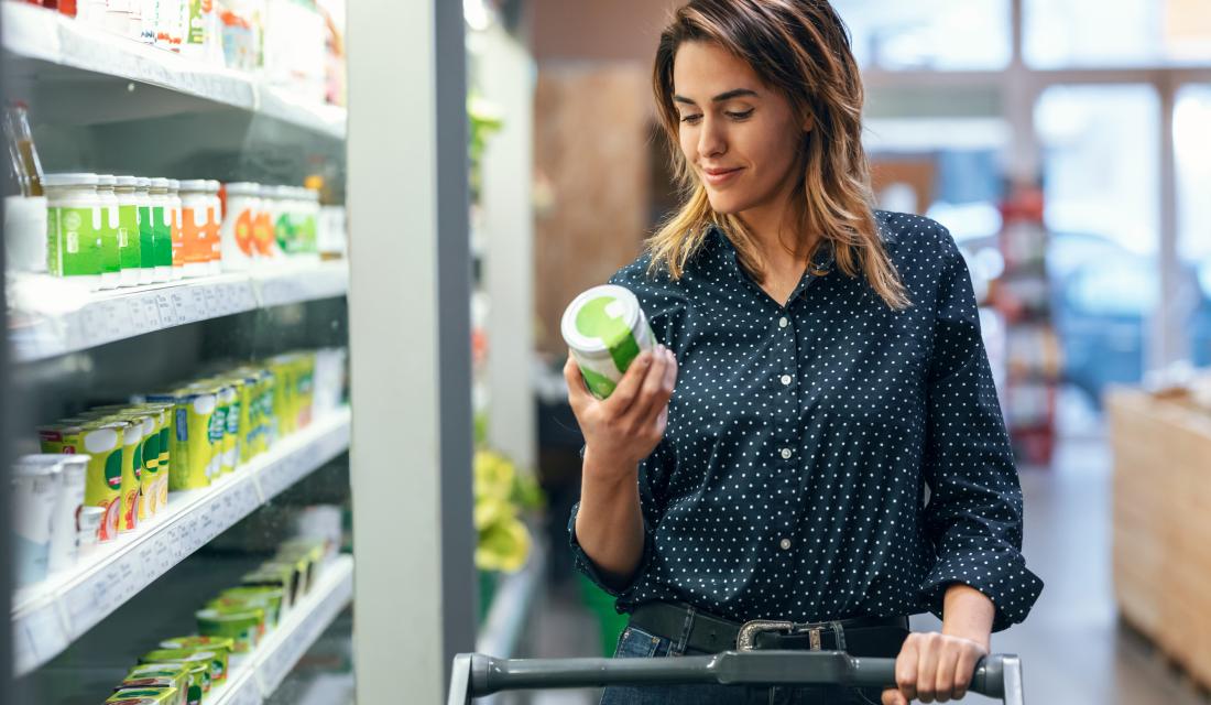 Frau mit Einkaufswagen im Supermarkt steht an der Kühltheke und liest sich die Rückseite eines Produkts durch 