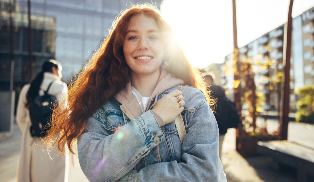 Eine junge rothaarige Frau lacht in die Kamera. Hinter ihr ist ein verglastes Gebäude und die Sonne scheint. Sie trägt einen Rucksack und im Hintergrund sieht man weitere Personen mit Rucksack.