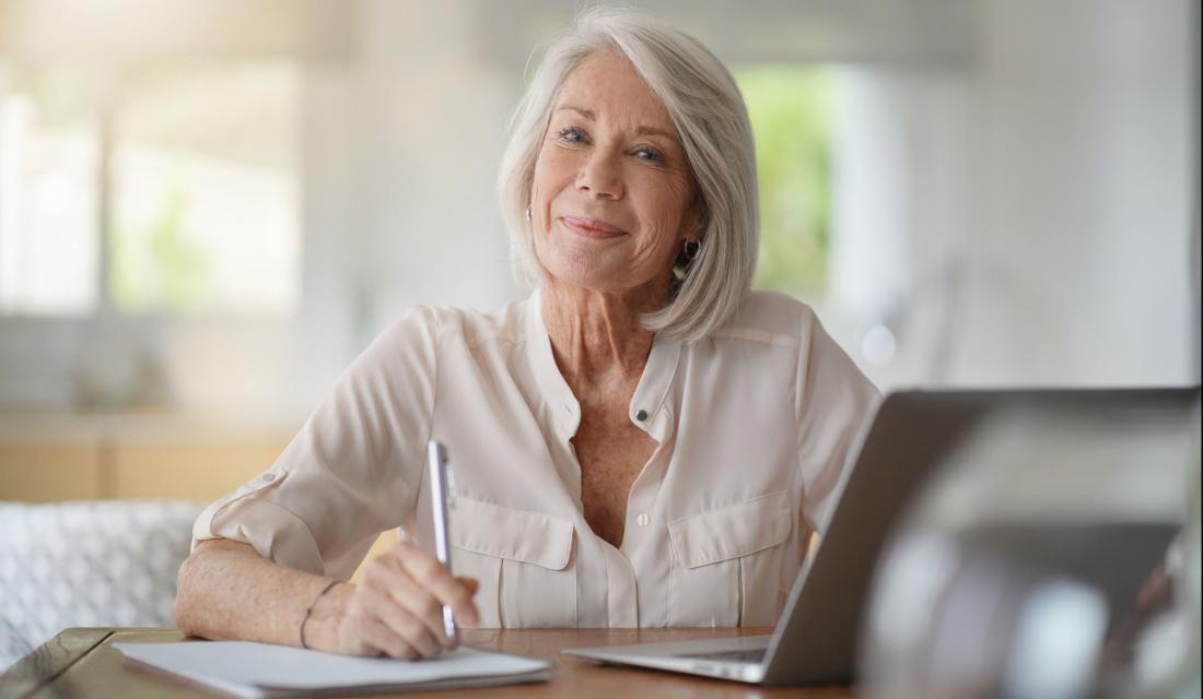 Eine Frau mit grauen kurzen Haaren sitzt am Tisch vor einem Laptop. In ihrer Hand hält sie einen Stift, vor ihr liegt ein Notizblock. Sie lächelt in die Kamera.
