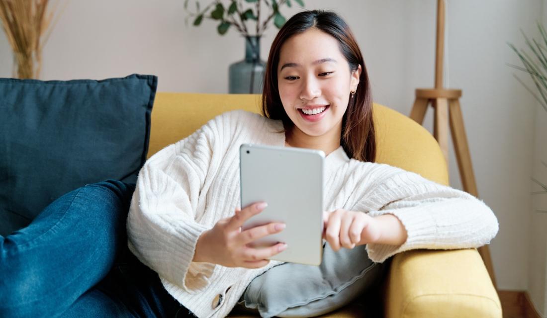 Eine junge Frau sitzt auf einem gelben Sofa im Wohnzimmer. In den Händen hält sie ein Tablet, schaut darauf und lacht.