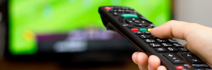 Foto von einer Hand, die eine Fernbedienung hält und auf einen Fernsehbildschirm richtet, auf dem ein Fußballspiel läuft