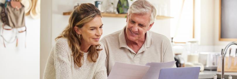 Mann und Frau mittleren Alters sitzen an der Küchentheke in einem lichtdurchfluteten Raum und schauen gemeinsam auf Unterlagen. Vor ihnen steht ein Laptop. Sie lächeln.