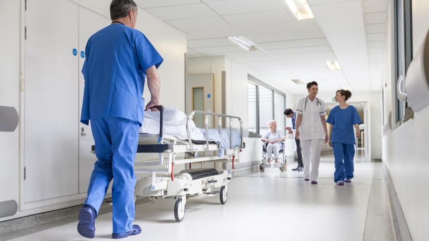 Szene auf einem Krankenhausflur: Ein Pfleger schiebt ein leeres Bett, ein Arzt spricht mit einer Frau im Rollstuhl, eine Ärztin und eine Pflegerin gehen vorbei.