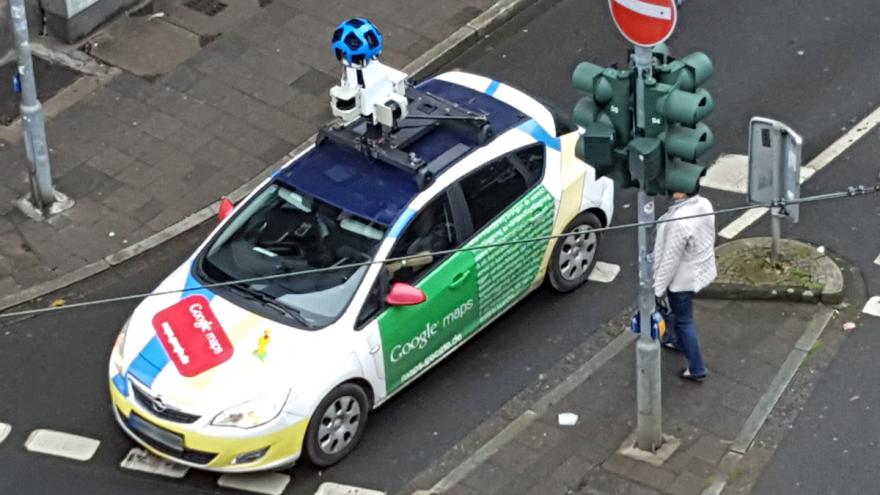 Ein Auto mit Google-Werbung und Rundum-Kamera auf dem Dach an einer Straßenkreuzung.