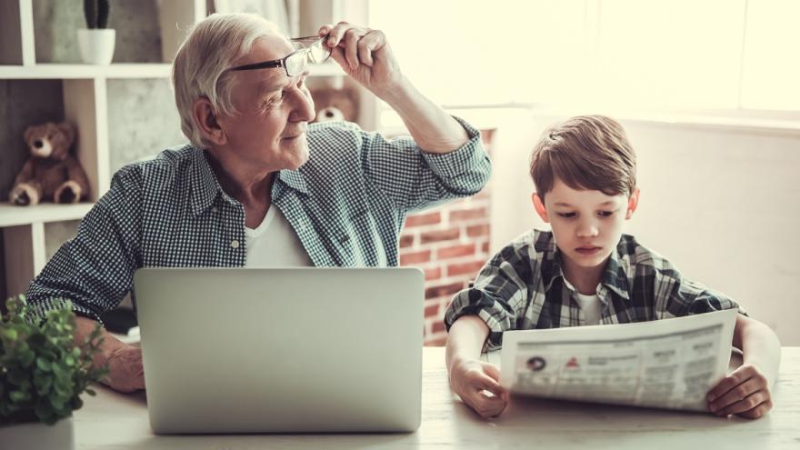 Älterer Mann am Computer. Neben ihm sitzt ein Junge und liest Zeitung.