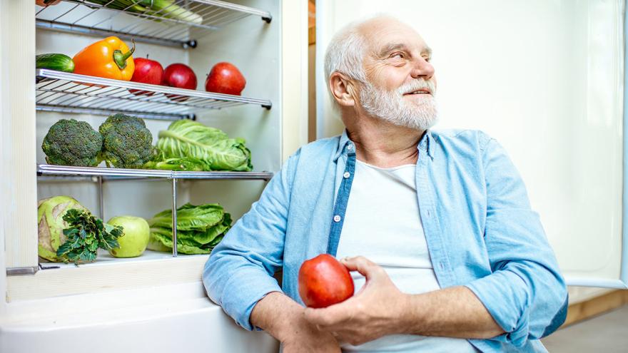 Senior sitzt am offenen Kühlschrank mit Obst und Gemüse