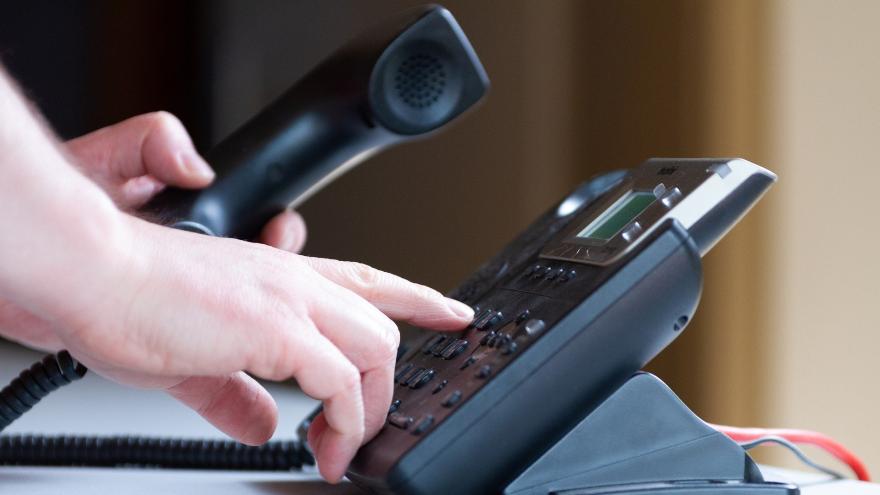 Eine Person hat den Telefonhörer eines Festnetztelefons in der linken Hand und wählt mit der rechten Hand eine Nummer.
