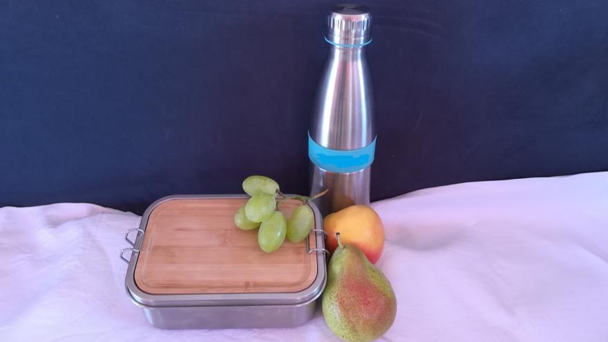 Auf einer Tischdecke stehen eine Brotdose, eine Trinkflasche und Obst.