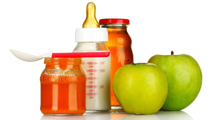 Im Vordergrund links steht ein Gläschen mit orange-rotem Babybrei. Darauf liegt ein Plastiklöffel für Babys. Rechts daneben liegen zwei grüne Äpfel. Im Hintergrund steht eine Babyflasche aus Glas, gefüllt mit Milch und eine kleine, verschlossene Glasflasche mit Apfelsaft.