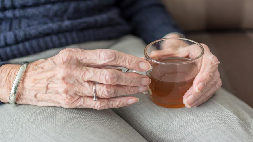 Die Hände einer älteren Dame halten ein Glas mit Schwarztee. Sie stellt das Glas auf ihrem Schoß ab. An ihrer Hand sieht man ein silbernes Armband und einen silbernen Ring mit kleinem weißen Stein. Ihre Hose ist hellgrau, ihr Pullover dunkelblau.