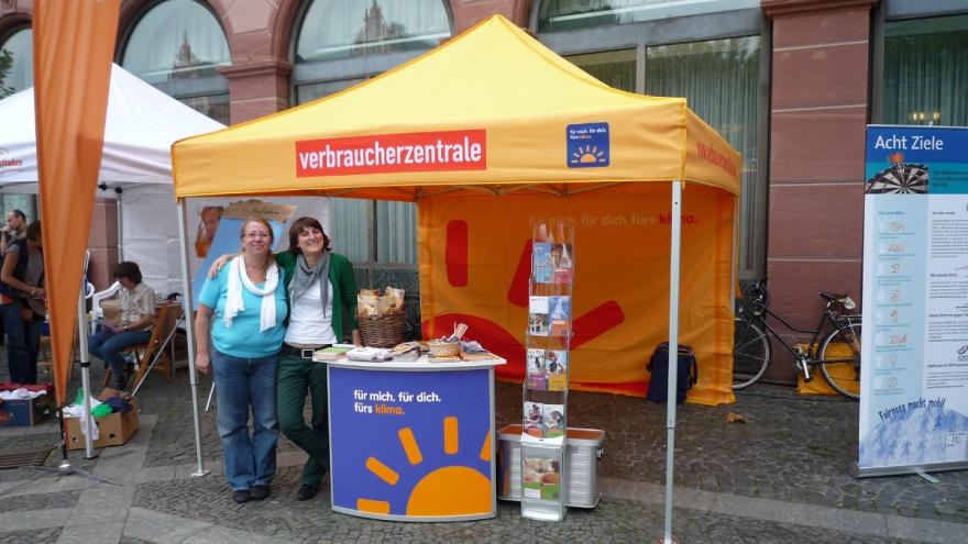 Zwei Frauen stehen an einem Straßenstand mit Infomaterial und blicken in die Kamera.