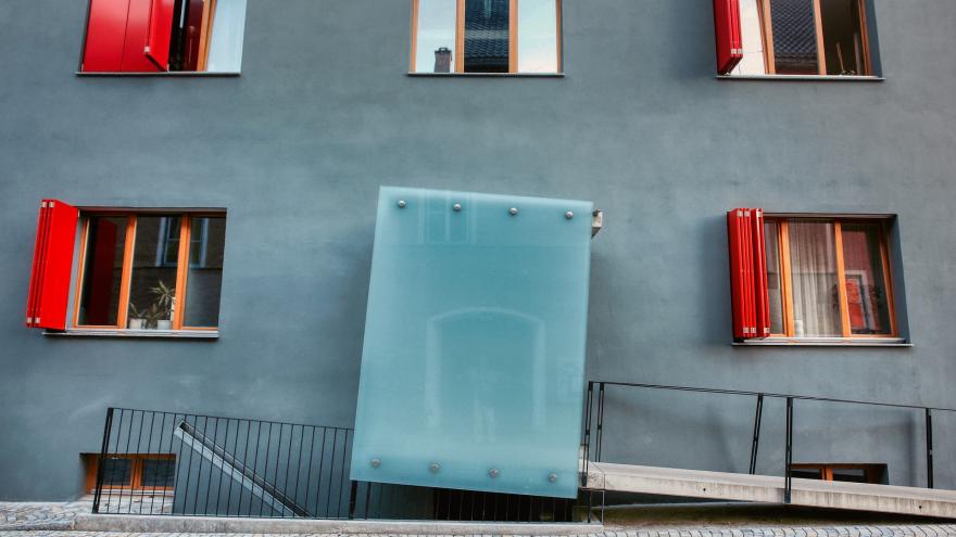 Eine Rampe führt zu einer Tür, die hinter einer Glasscheibe zu sehen ist. Um die Tür herum befinden sich unterschiedlich geöffnete Fenster von Wohnungen.