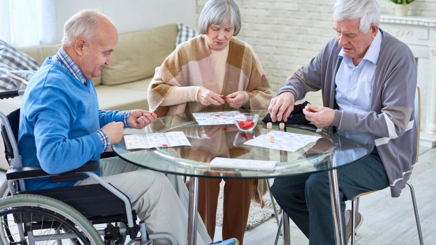 2 ältere Männer, einer davon im Rollstuhl, und eine ältere Frau sitzen am Tisch und spielen Karten