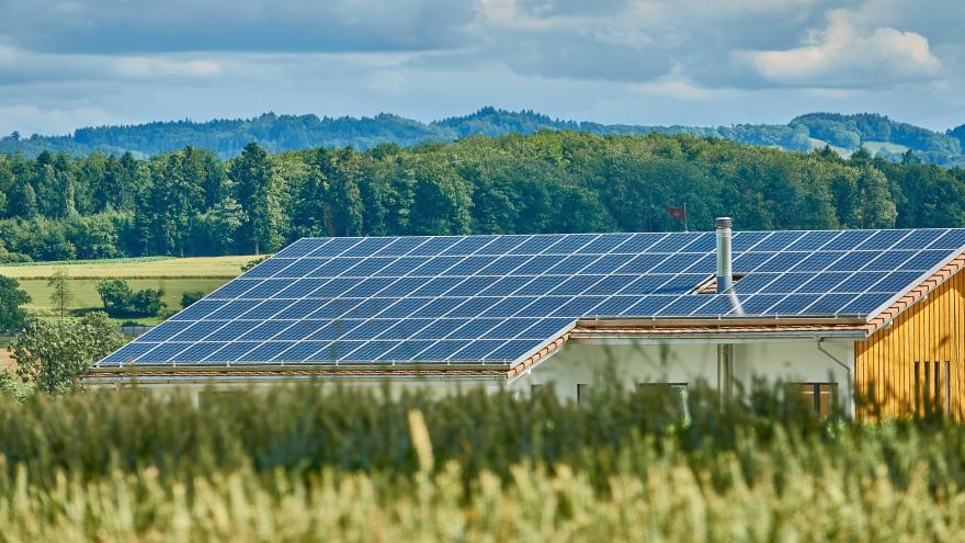 In mitten einer Landschaft in verschiedenen Grüntönen ist ein Hausdach mit Schornstein zu sehen. Das Dach ist komplett mit Solarpanels einer Photovoltaikanlage bedeckt.