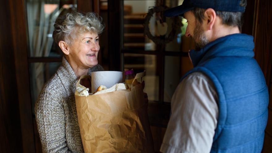Ältere Frau steht an der Haustür und bekommt eine Tüte mit Lebensmitteln von einem jungen Mann überreicht.