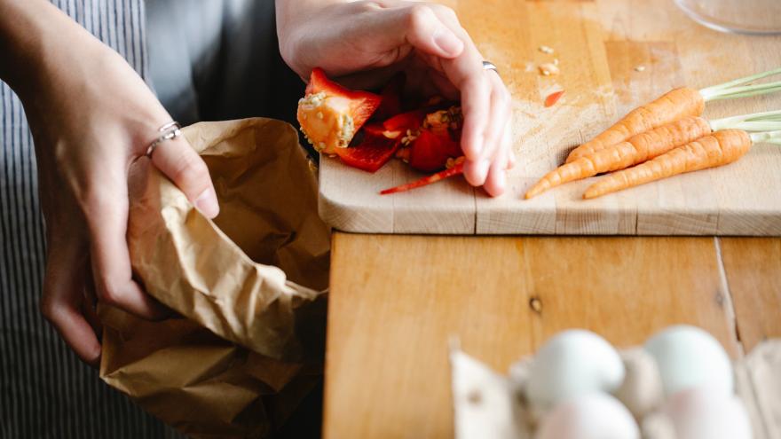 Eine Frau steht in der Küche. Der Bildausschnitt zeigt ihre Hände. In der linken Hand hält sie eine Papiertüte und mit der rechten Hand schiebt sie Gemüsereste von der Arbeitsfläche Richtung Tüte. Vorne dran steht ein Karton mit Eiern. 