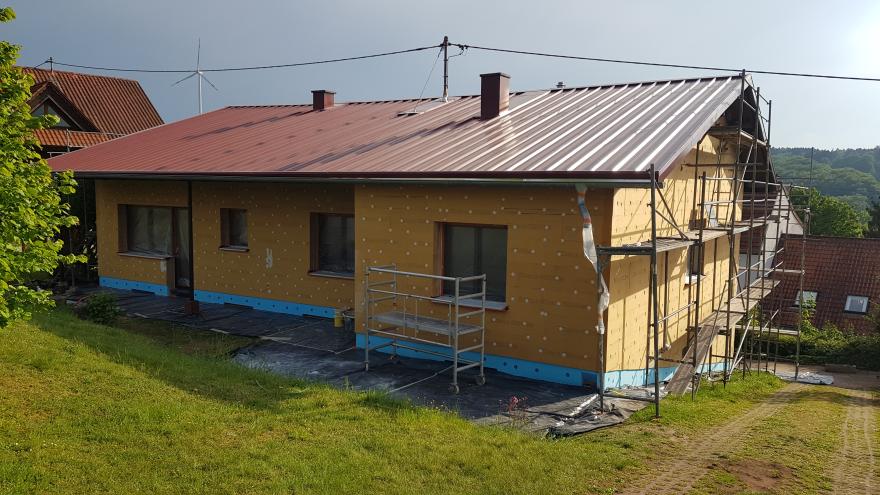 Ein einstöckiges Haus wird saniert. Wärmedämmplatten werden außen am Haus angebracht. Verschiedene Baugerüste sind zu sehen.