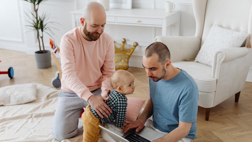 Zwei Männer sitzen auf dem Boden. Einer der Männer hält einen Säugling an der Hüfte, der andere hat einen Laptop auf dem Schoß.