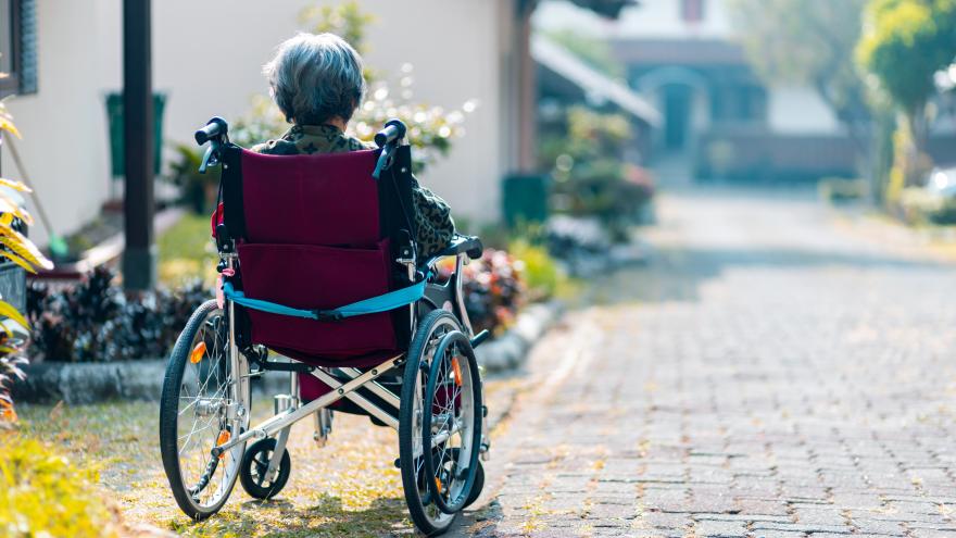 Eine ältere Frau sitzt im Rollstuhl. Der Rollstuhl steht am Rand einer ruhigen Straße in einer Wohnsiedlung.