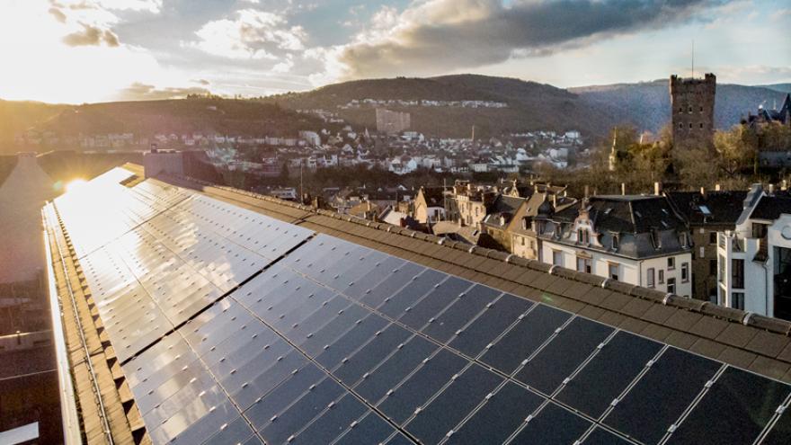 Draufsicht. Eine lange Dachfläche ist mit Solarpanels einer Photovoltaik-Anlage bedeckt. Die Sonne scheint von links auf die Panels. Im Hintergrund ist das Panorama der Stadt Bingen zu erkennen.