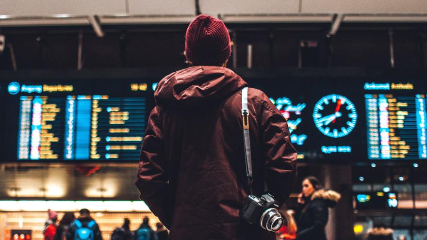 Ein junger Mann steht vor einer Reisetafel am Flughafen. Er ist von hinten zu sehen und hat eine Fotokamera umhängen.