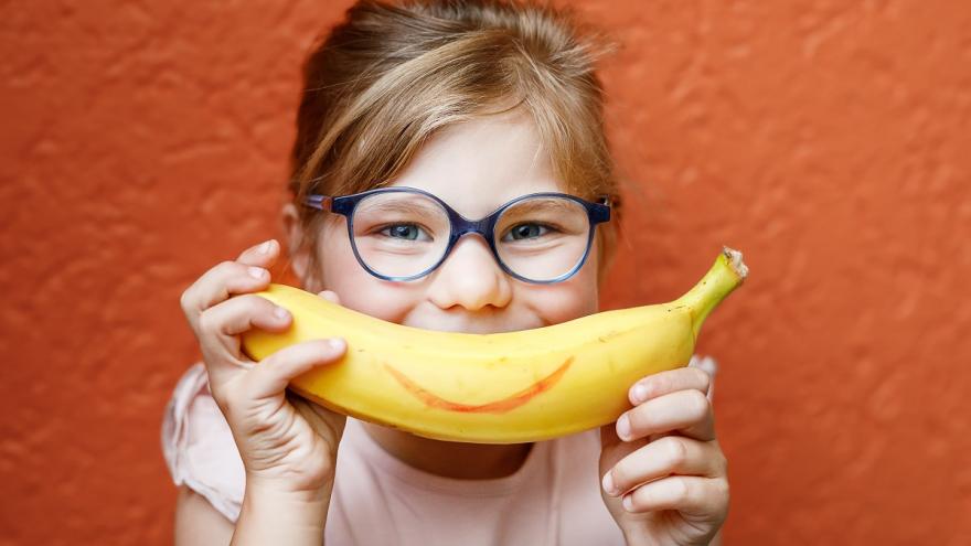 Ein Mädchen mit Brille hält sich einen Banane mit einem aufgemalten Lächeln vor den Mund. 