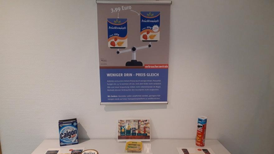 Ein Plakat an der Wand. Davor ein Tisch mit verschiedenen lebensmittelverpackungen.