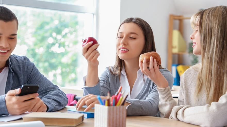 Drei Jugendliche sitzen im Klassenraum, unterhalten sich und haben ein Handy, einen Apfel und ein Hefestückchen in der Hand.