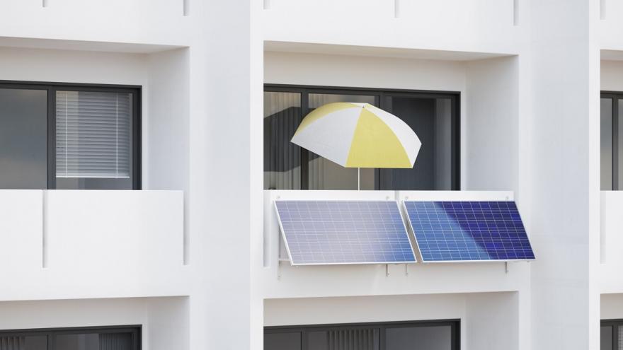 Ein Mehrfamilienhaus. An einem Balkon hängt ein Balkonkraftwerk und steht ein Sonnenschirm.