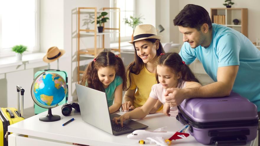 Eine Familie am Laptop. Daneben steht ein Reisekoffer.