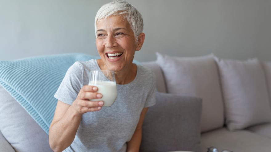 Lachende Frau mit grauen Haaren sitzt auf der Couch mit einem Glas Milch in der Hand 