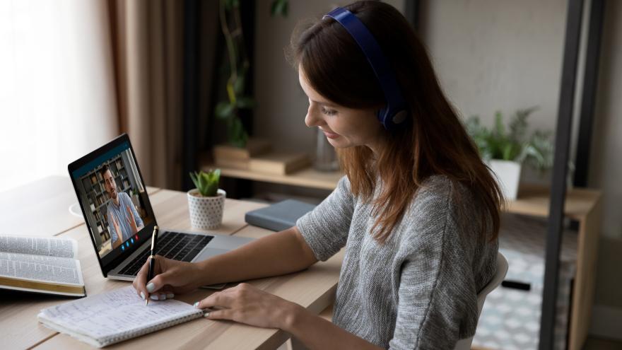 Frau sitzt am Schreibtisch vor Laptop und hat ein Videogespräch mit einem Mann. Sie schreibt Notizen auf einen Block. Es sieht aus wie ein Coachinggespräch.