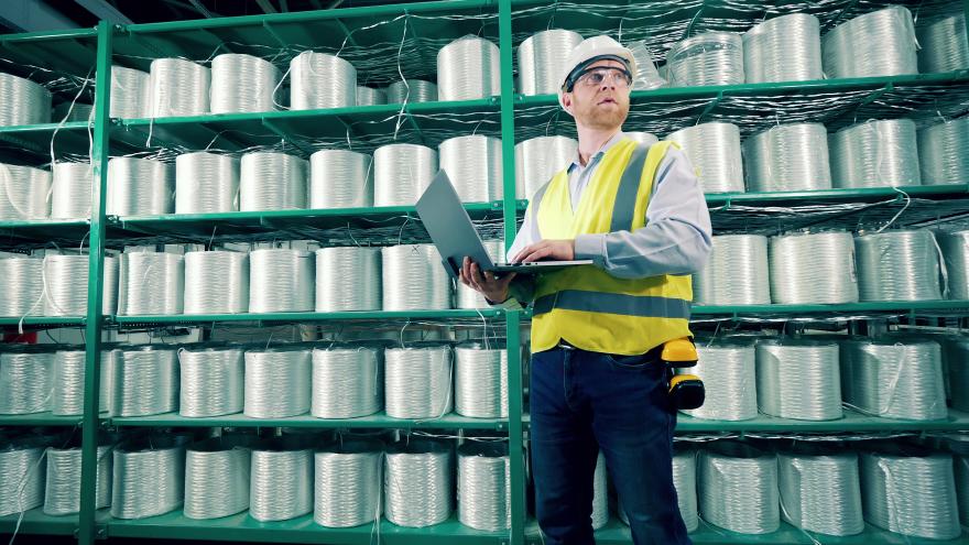 Techniker mit Warnweste und Helm und Laptop in der Hand steht vor einem großen Regal mit Glasfaserrollen.