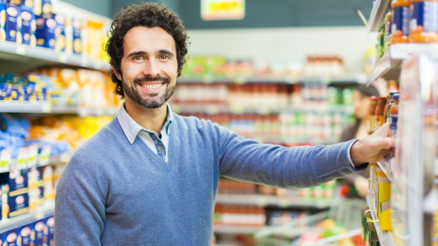 Ein Mann mit braunen Locken und Bart steht im Supermarkt und greift in ein Regal nach einem Produkt. Er lacht freundlich in die Kamera.