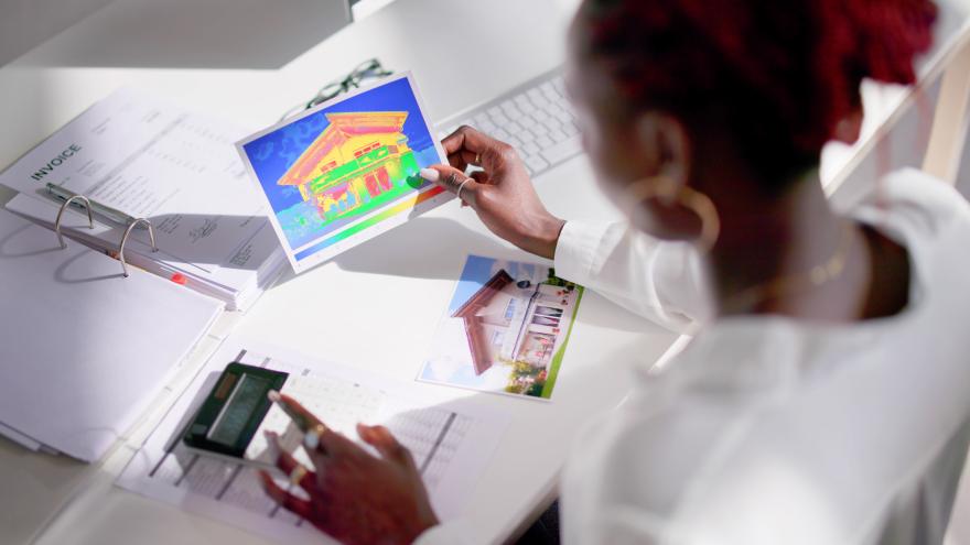 Eine Frau sitzt am Schreibtisch und wird über ihre Schulter fotografiert. Sie hält ein Wärmebild eines Hauses in der Hand. Vor ihr liegt ein Ordner und Unterlagen sowie ein Taschenrechner.