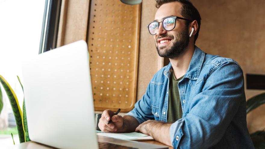 Junger Mann mit Brille sitzt in einem Café vor dem Laptop, er hält einen Stift in der rechten Hand und schreibt in einen Notizblock. Er blickt aus dem Fenster und lacht.